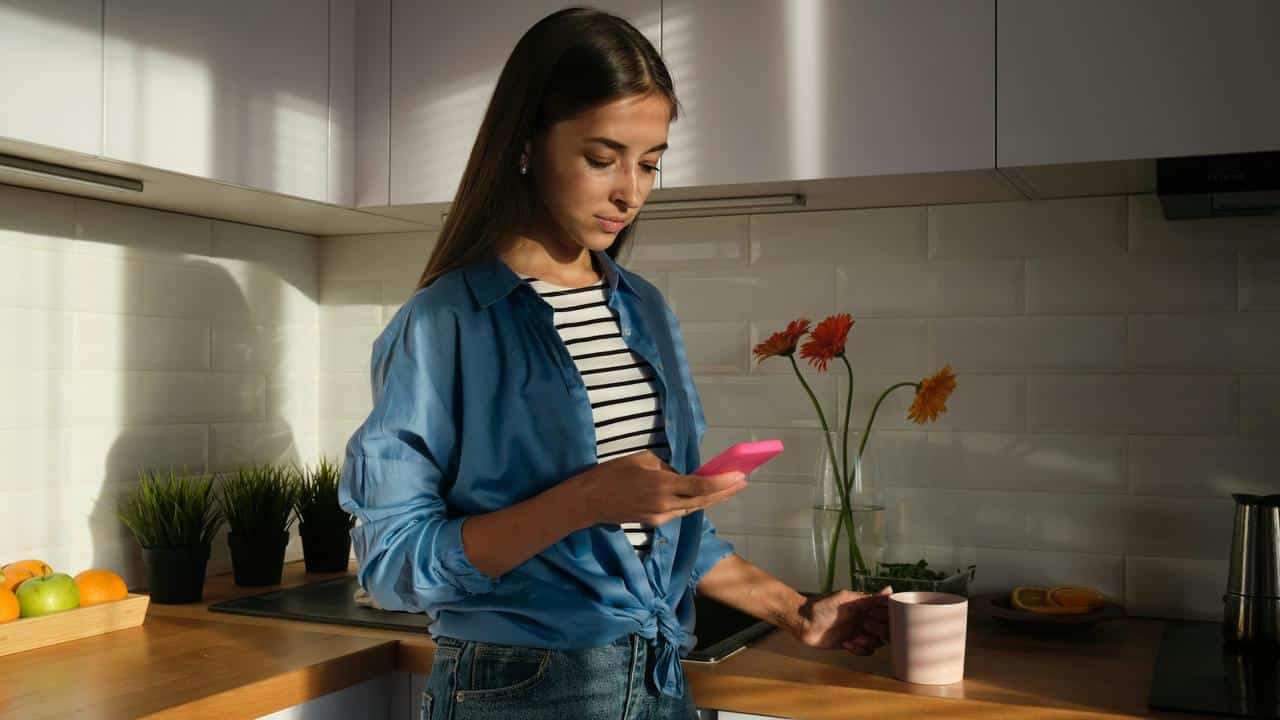 A young woman in a kitchen looks at her phone while holding a mug.