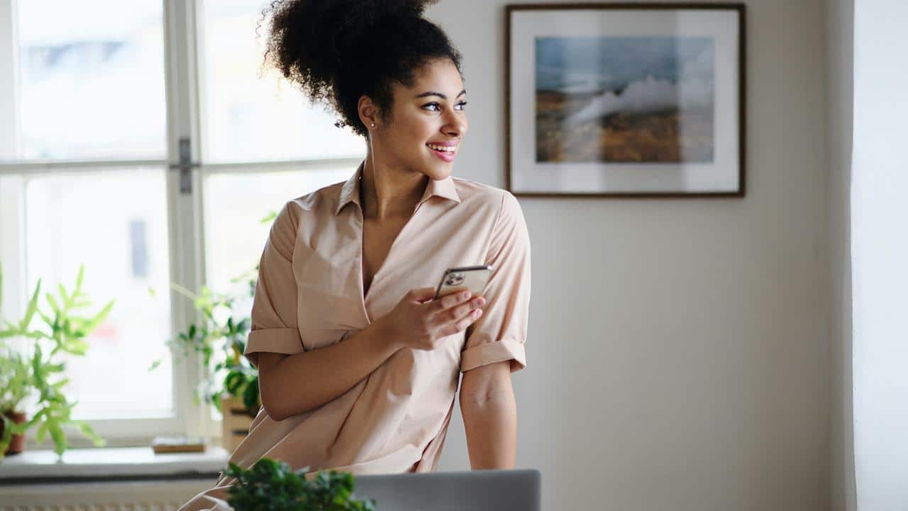 A smiling woman holding a smartphone looks out of a window while sitting on a desk.
