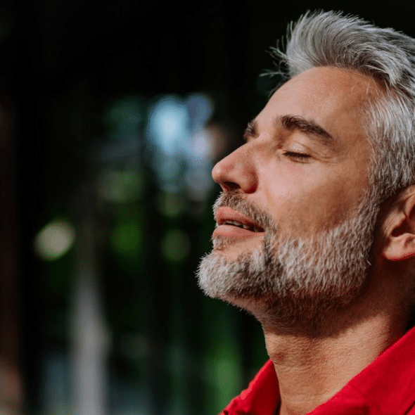 A relaxed, gray-haired man with a beard stands outdoors with his eyes closed, and his face turned to the side.