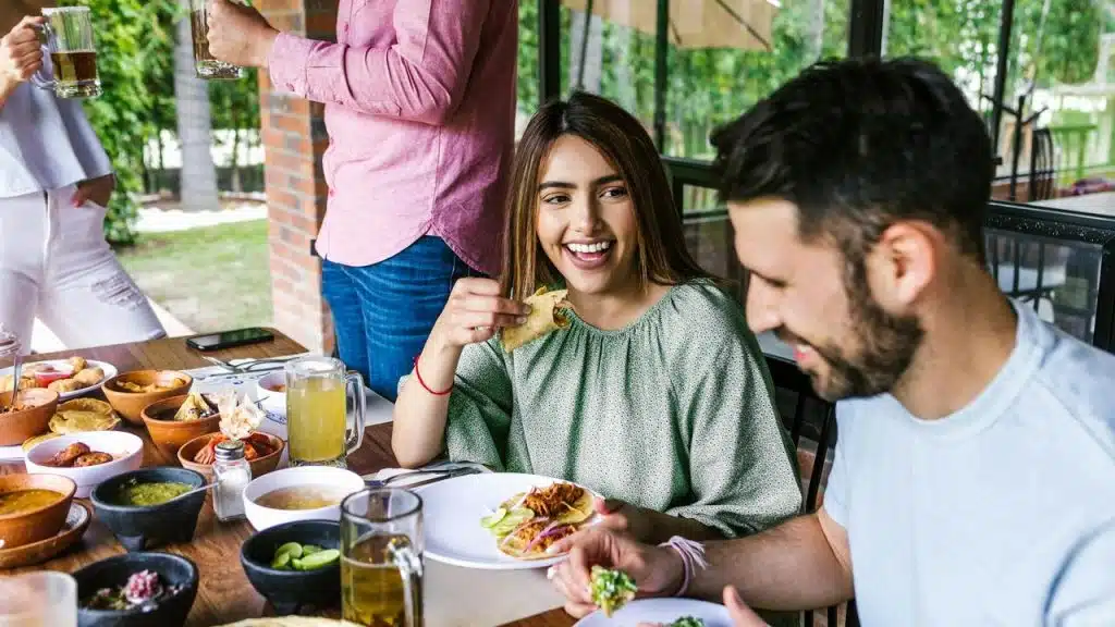 A woman and man enjoy tacos and drinks while laughing at a lively outdoor meal.