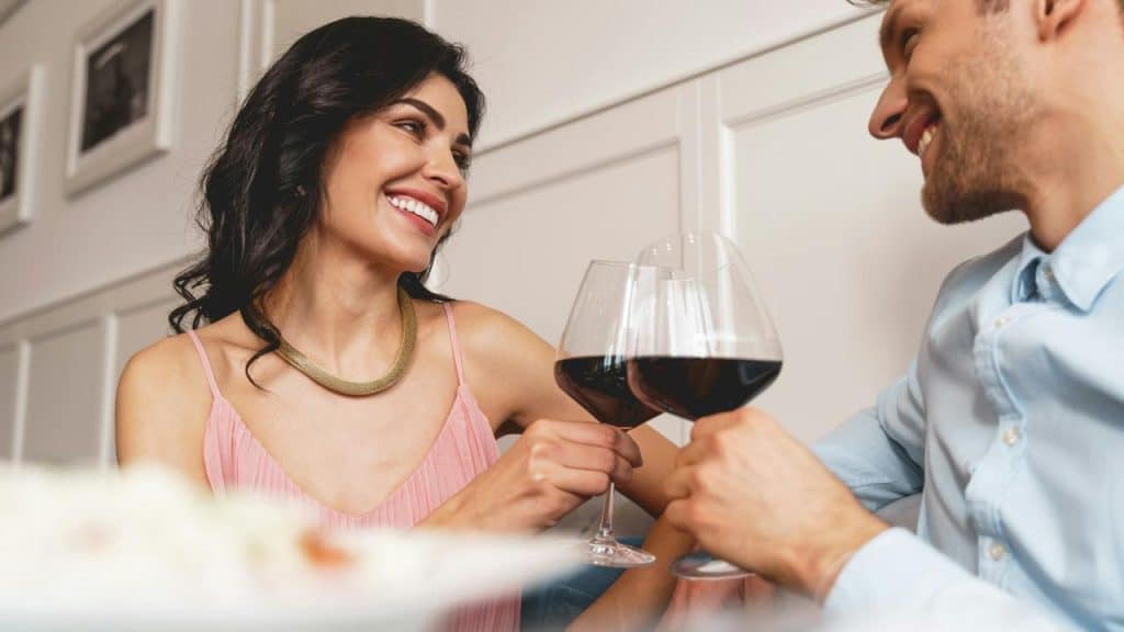 A smiling couple clinks glasses of red wine while enjoying a romantic moment together.