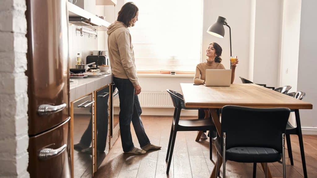 A man standing in a kitchen while a woman sits at the table with a laptop and a glass of juice.