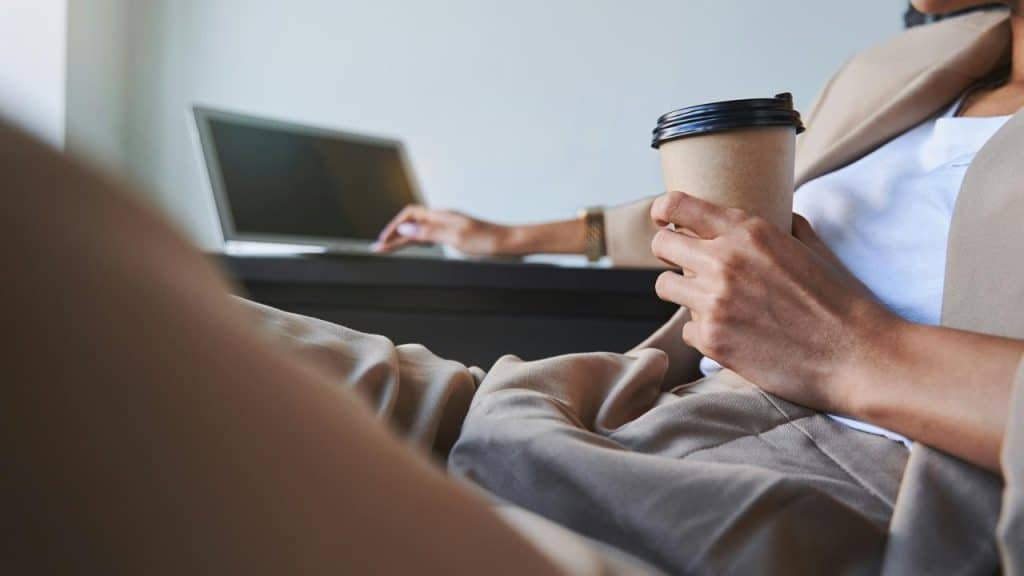 A person in business attire holding a coffee cup while working on a laptop.