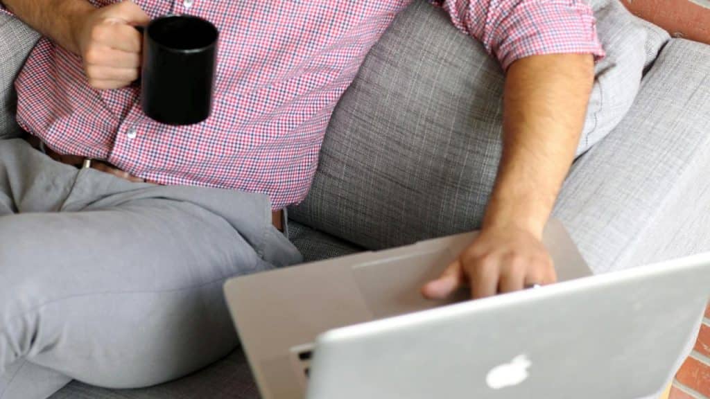A man sitting on a couch holding a coffee mug while using a laptop.