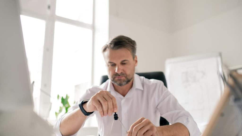 A man in a white shirt working at a desk with a compass in hand.