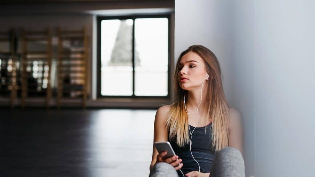 A young woman sitting on the floor with earphones in, holding a smartphone.