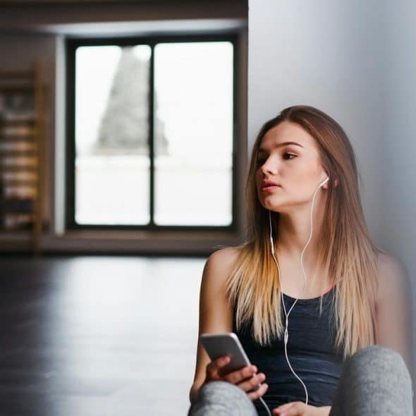 A young woman sitting on the floor with earphones in, holding a smartphone.