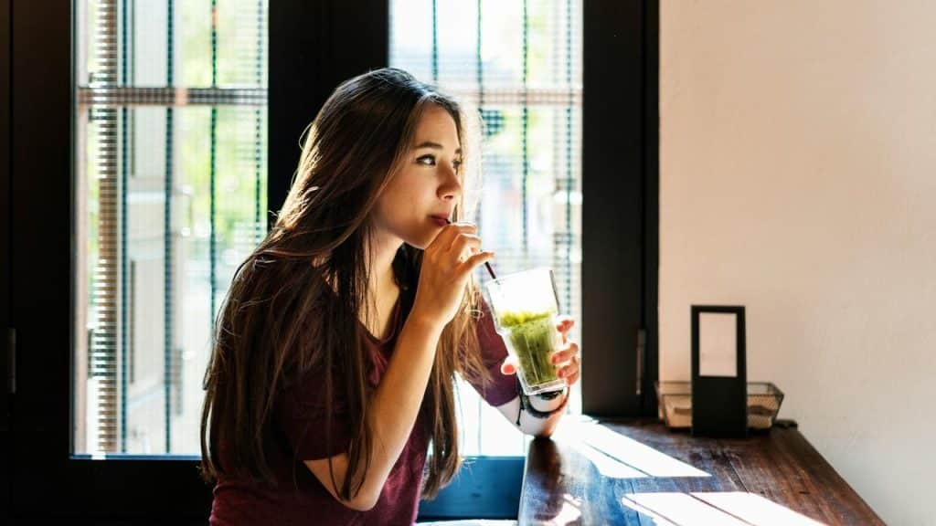 A young woman sitting by a window, drinking a green smoothie with a straw.