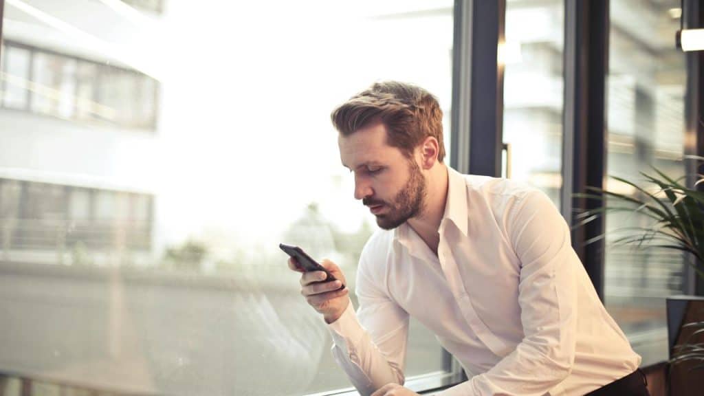 A man in a white shirt sitting by a window, looking at his phone.