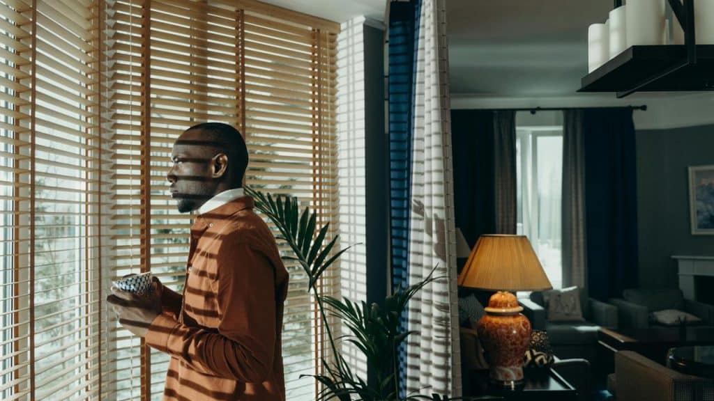 A man standing by a window with blinds, holding a cup and looking outside.