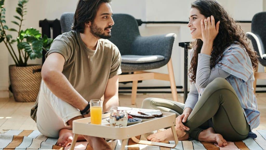 A man and a woman sitting on the floor and talking over breakfast.