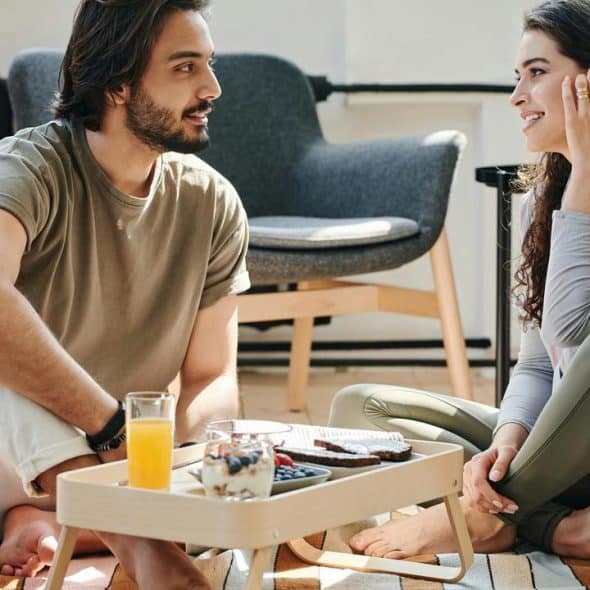 A man and a woman sitting on the floor and talking over breakfast.