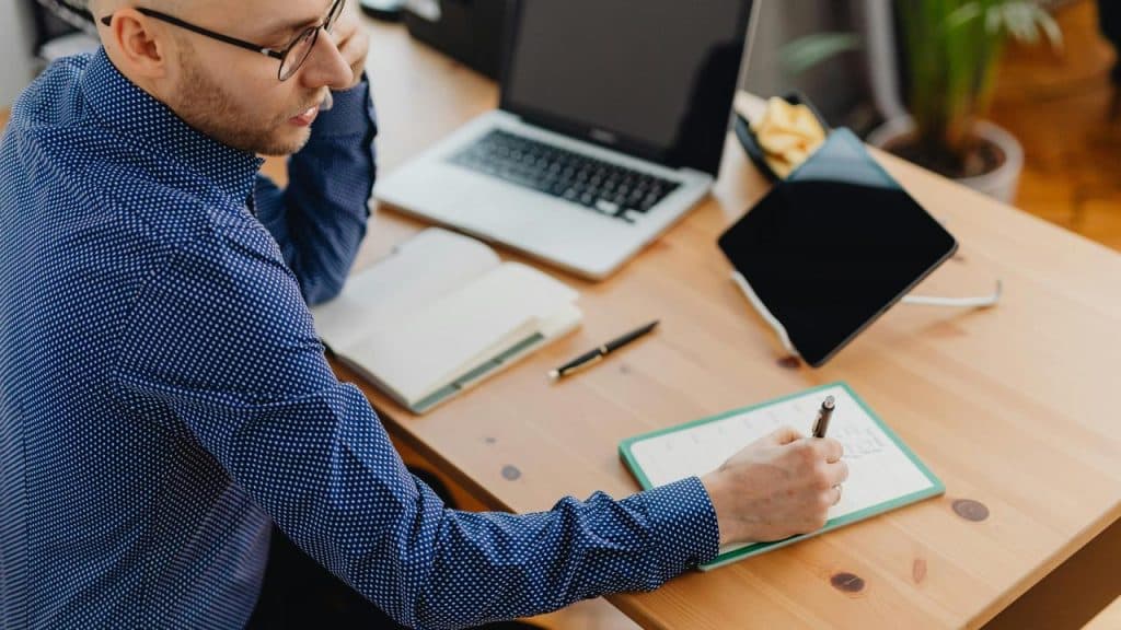 A man sitting at a desk writing in a notebook while on the phone.