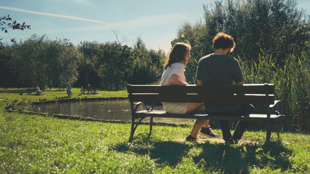 A man and a woman sitting on a bench by a pond in a park.