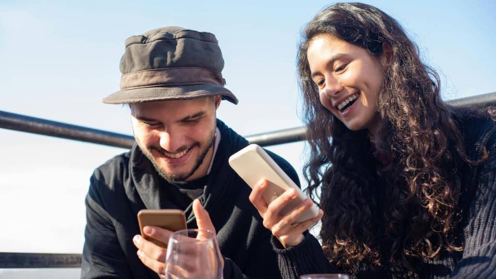 A man and a woman smiling while looking at their phones together.