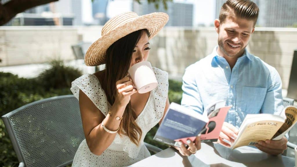 A woman drinking from a mug while a man reads a book beside her outdoors.
