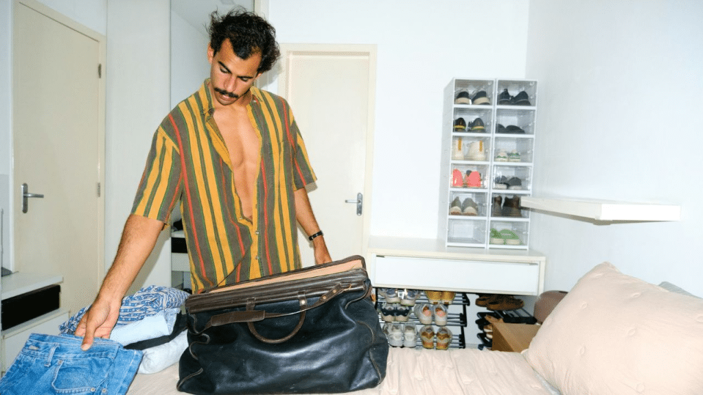 A man with an unbuttoned shirt and a mustache is packing a duffel bag on a bed.