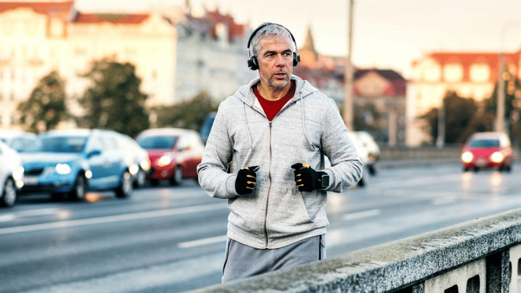 A gray-haired man wearing headphones and a hoodie is running on a city street.