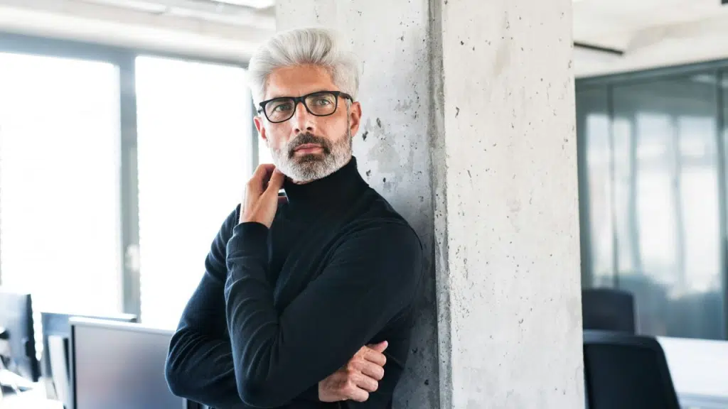 A man with gray hair, a beard, and glasses, wearing a black turtleneck, is looking directly at the camera with a serious expression.