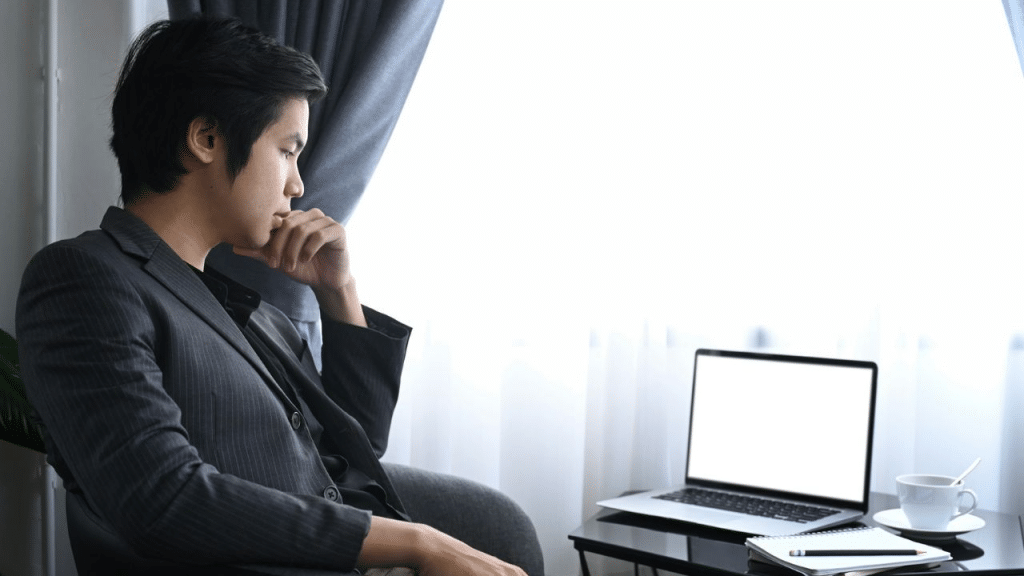 A serious man sits and stares thoughtfully at a laptop with a blank screen.