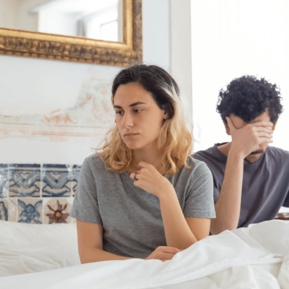 A man and a woman sit in bed. The man covers his face with his hand.