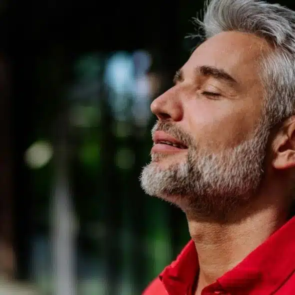 A handsome, gray-haired man in a red shirt stands with eyes closed and face turned toward the sky.