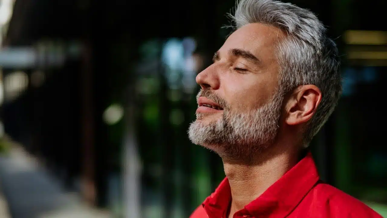 A handsome, gray-haired man in a red shirt stands with eyes closed and face turned toward the sky.