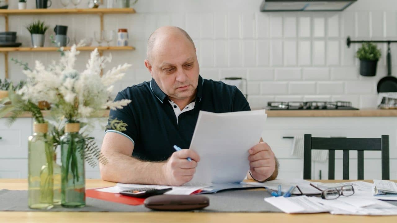A serious man in a black polo shirt reviews documents at a kitchen table.