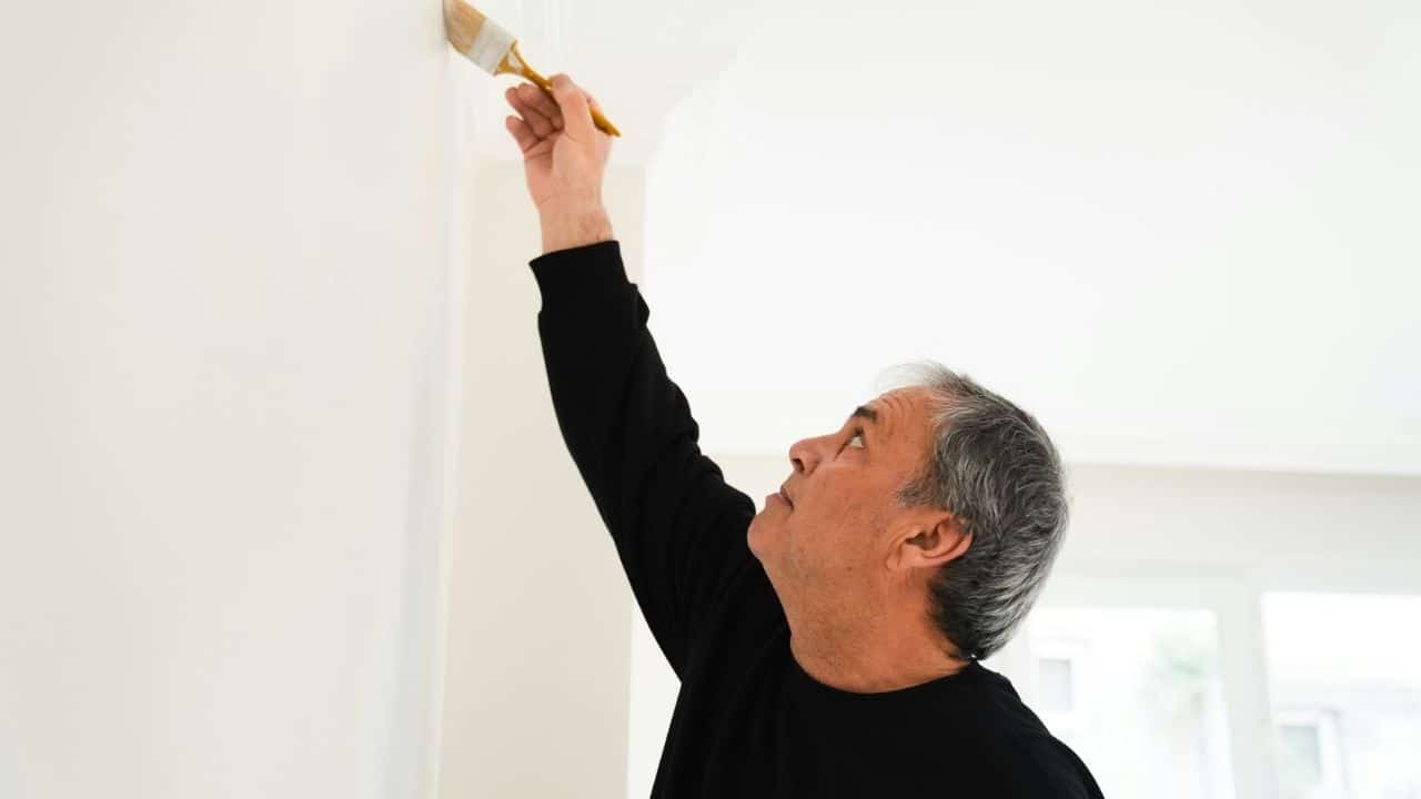 A gray-haired man in a black shirt painting a white wall.
