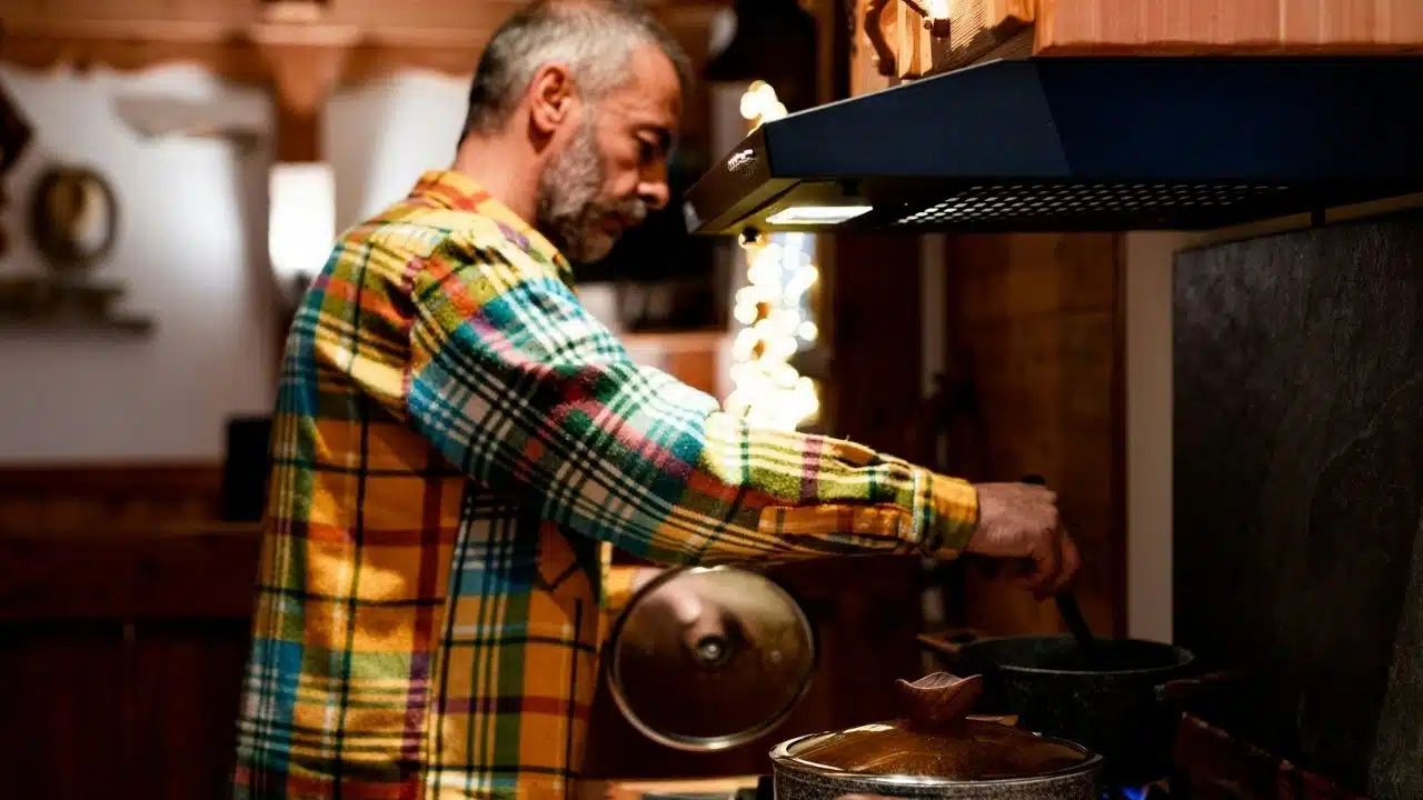 A focused, middle-aged man in a colorful plaid shirt stirs a pot in a kitchen.