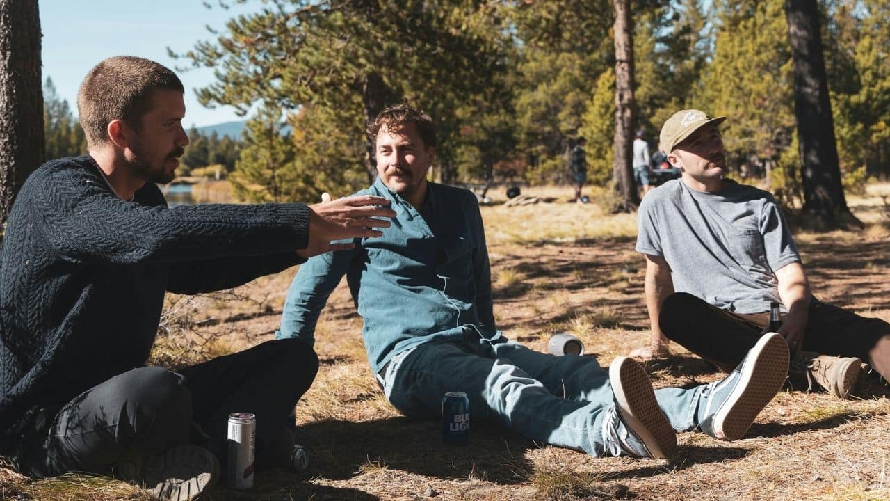Three friends sit and talk on the ground outdoors near a lake.