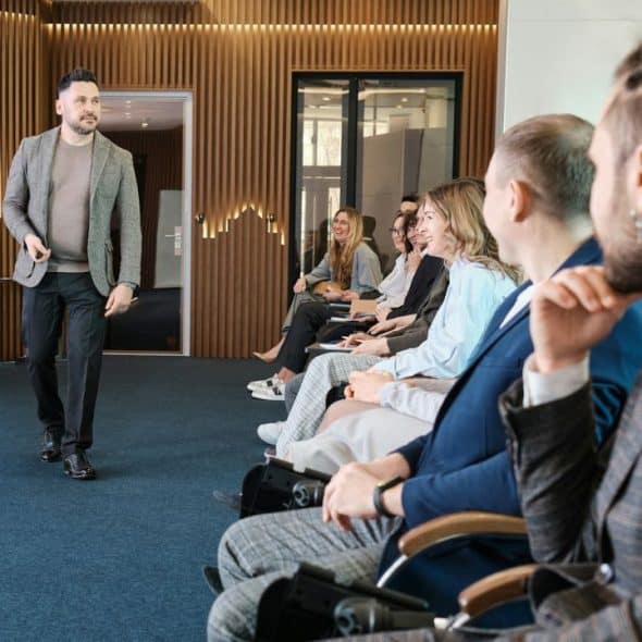 A speaker in a gray blazer is giving a presentation to a seated audience.