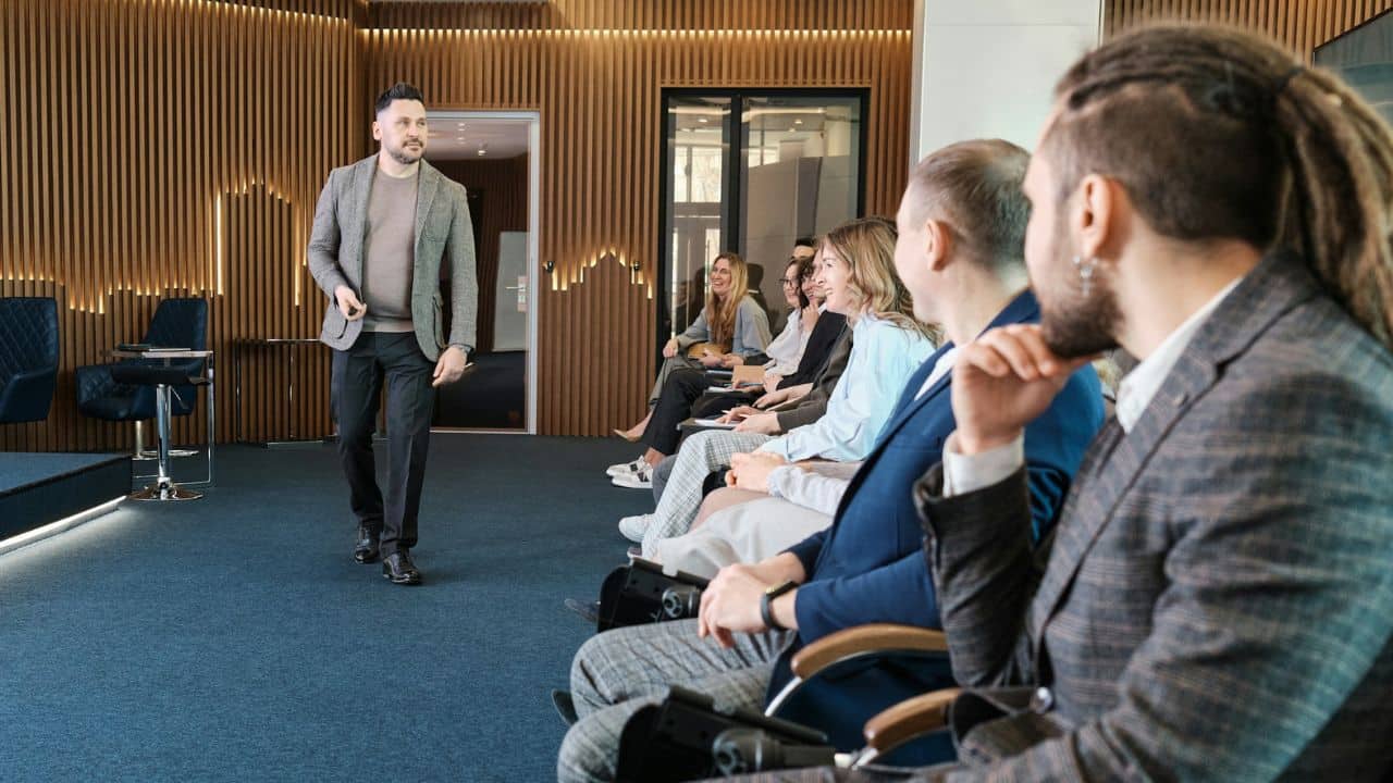 A speaker in a gray blazer is giving a presentation to a seated audience.