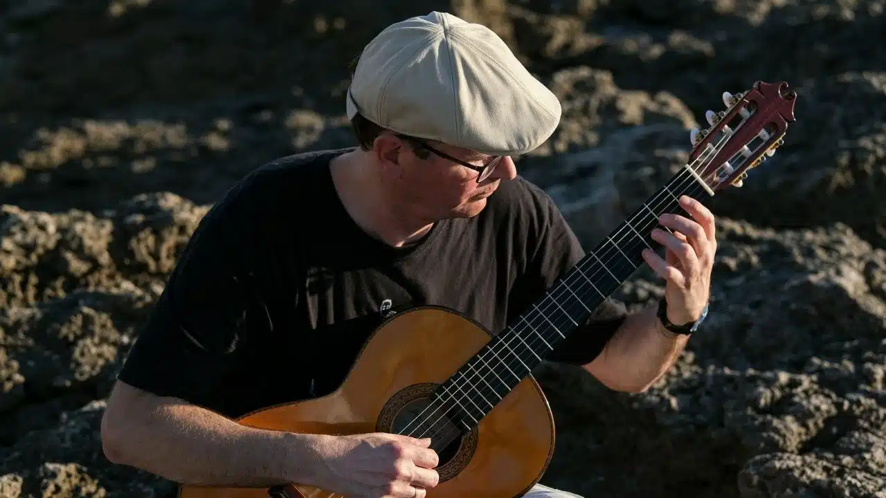 A person wearing a flat cap and glasses plays an acoustic guitar.