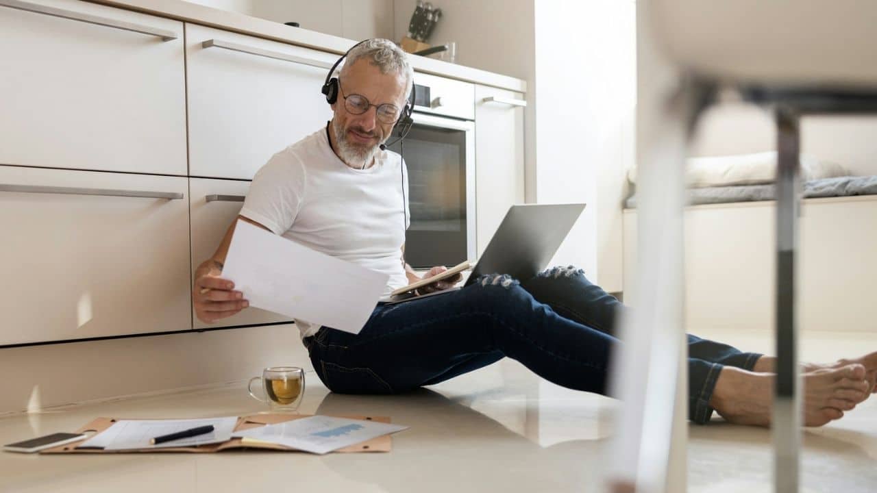 A mature man sits on a kitchen floor and reviews documents while using a laptop.