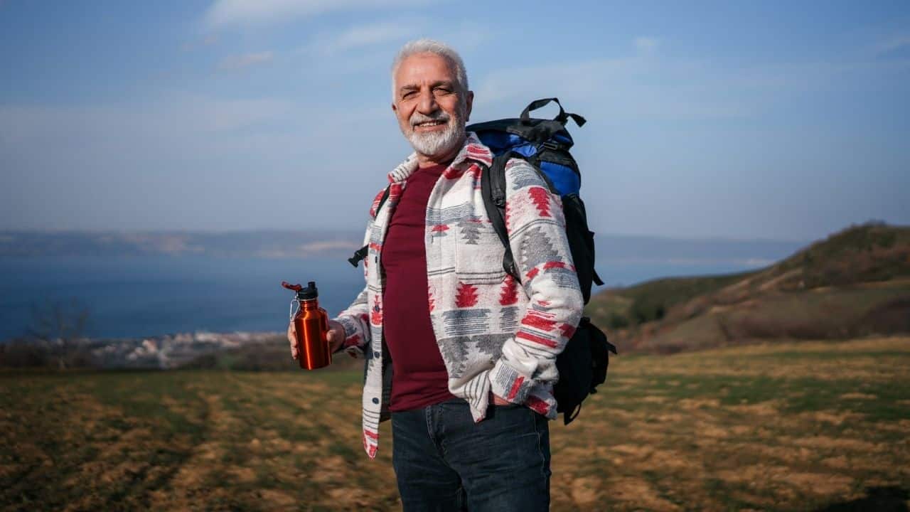 A senior man with a backpack and a water bottle smiles while hiking.