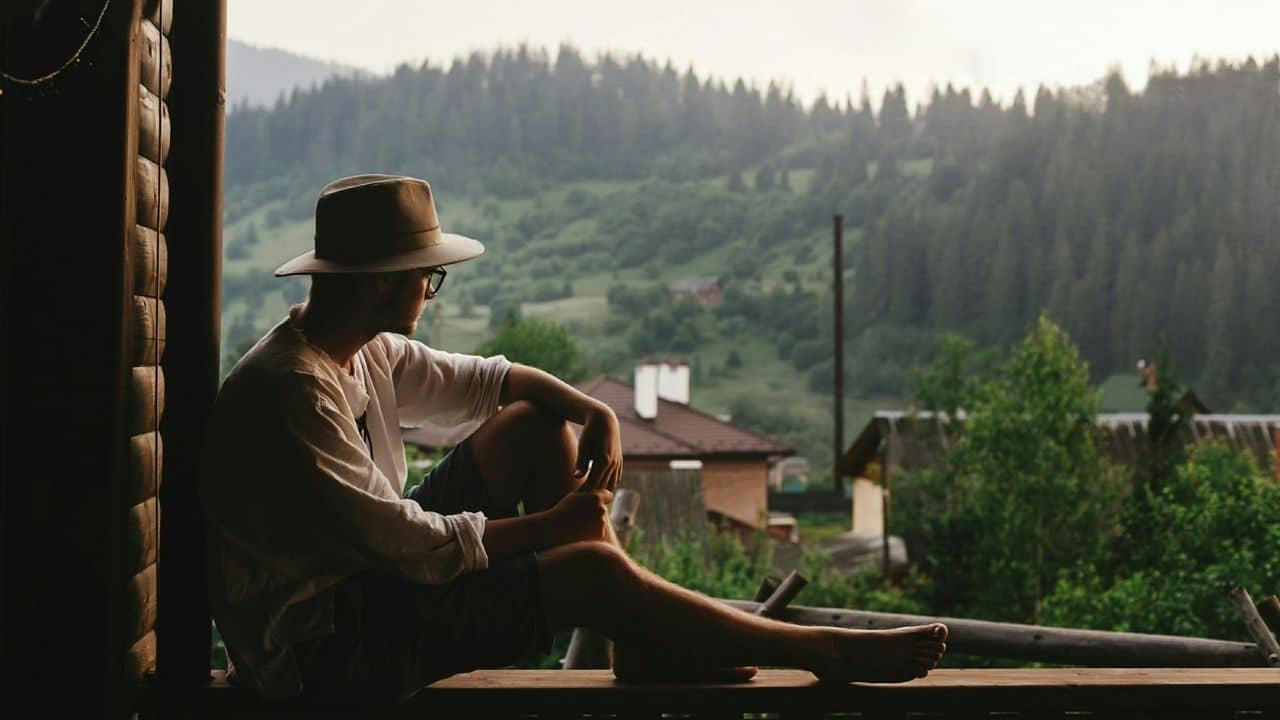 A barefoot man with a hat sits on a porch and looks out at a mountain range.