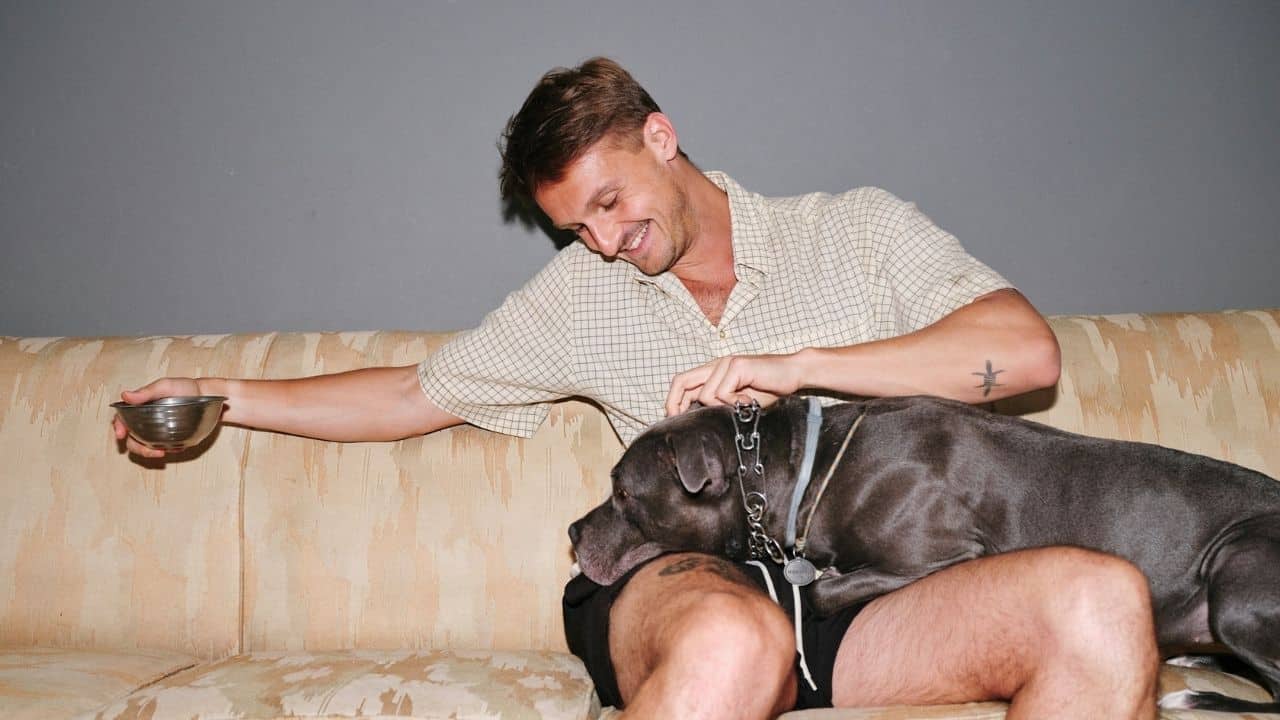 A joyful man sits on a couch with a gray dog and holds a bowl.