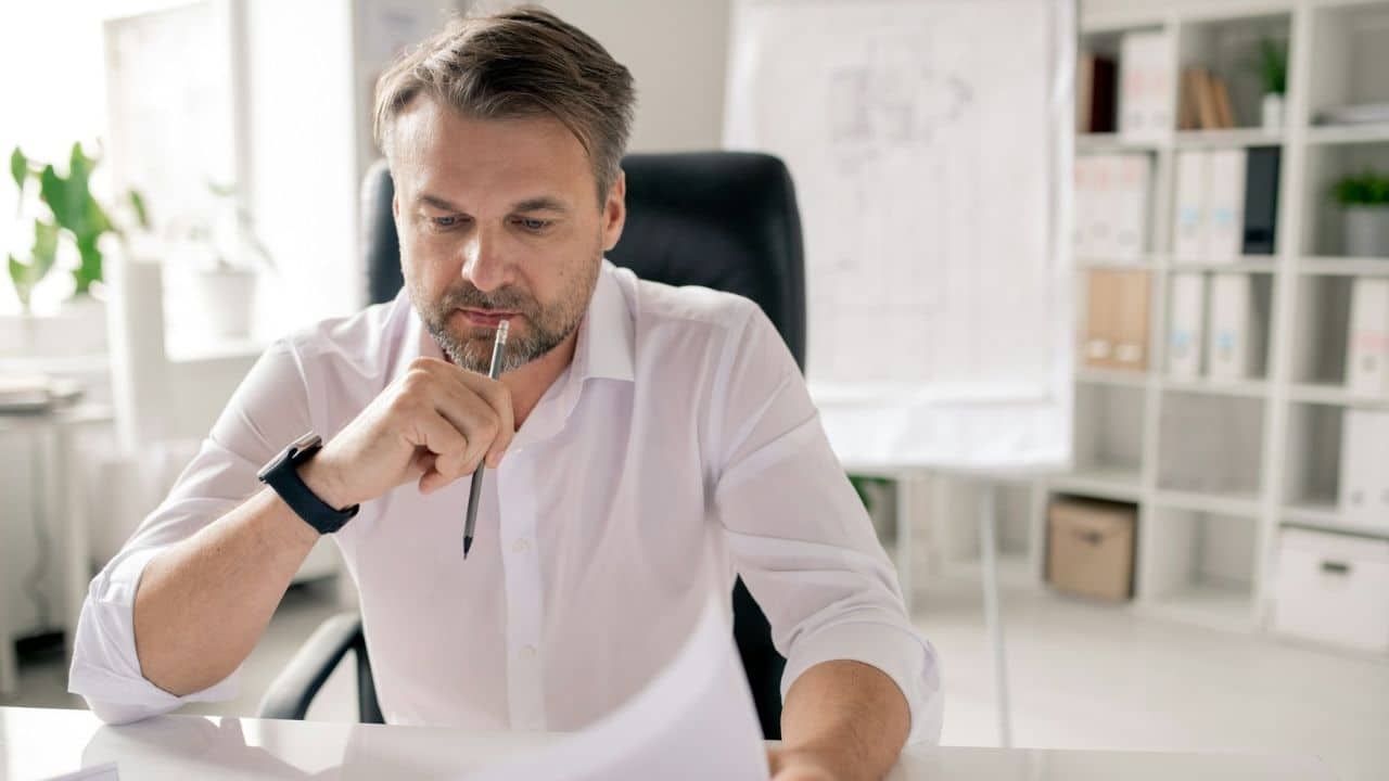 A middle-aged businessman with a beard and a white shirt looks at papers in an office.