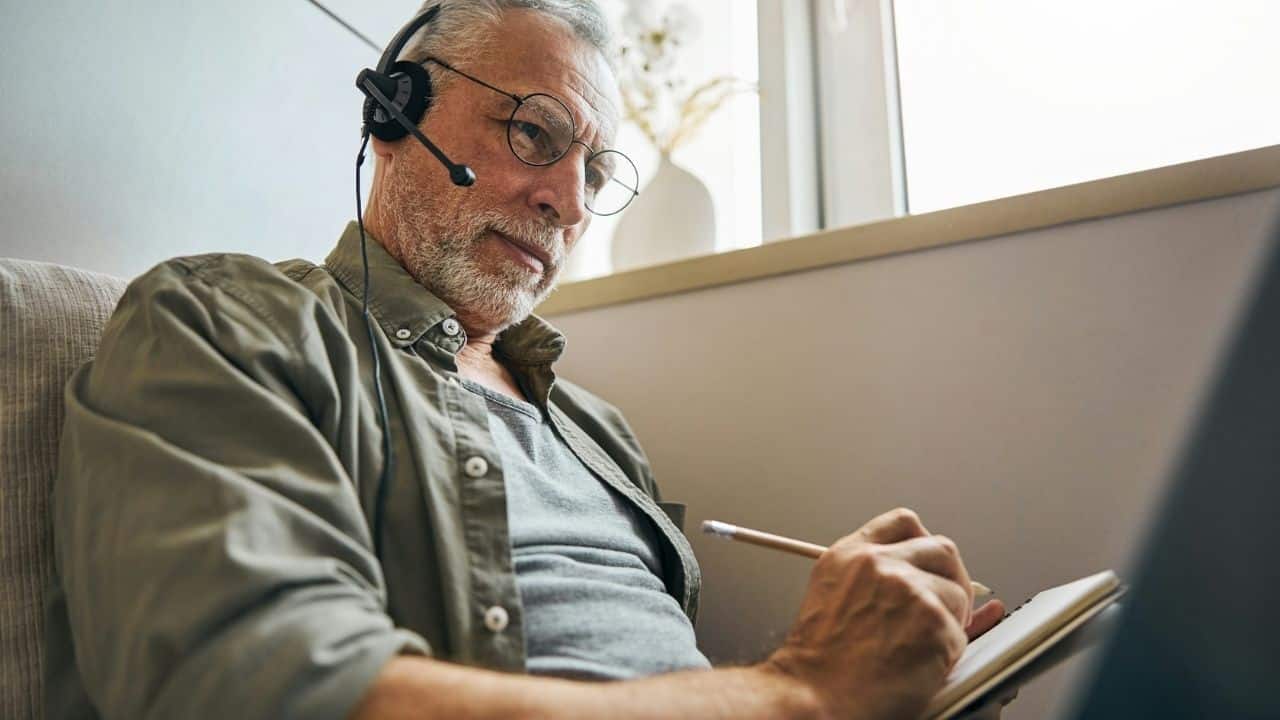 A serious senior man with a beard and headset writes in a notebook.