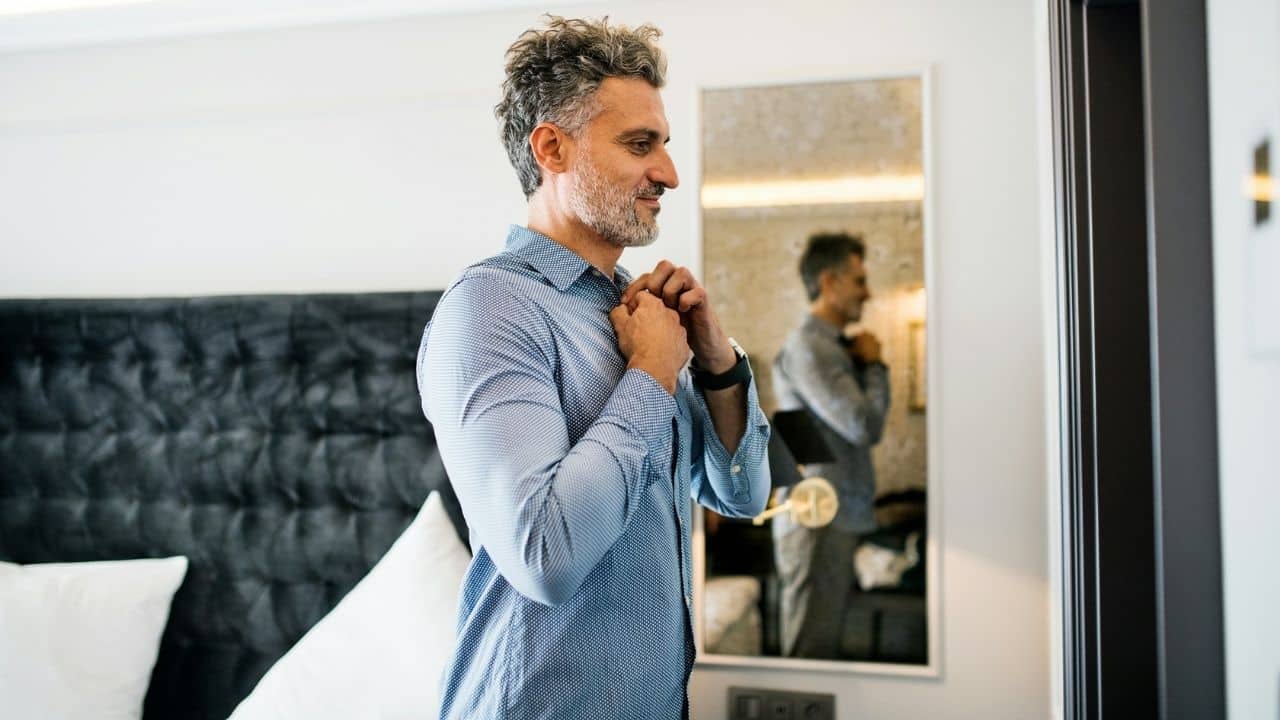 A smiling, gray-haired man in a blue shirt buttoning his collar in front of a mirror.