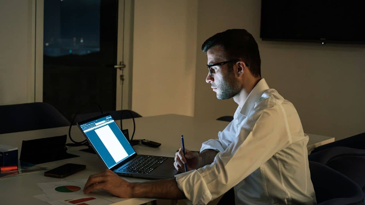 A determined businessman with glasses is working on his laptop late at night.