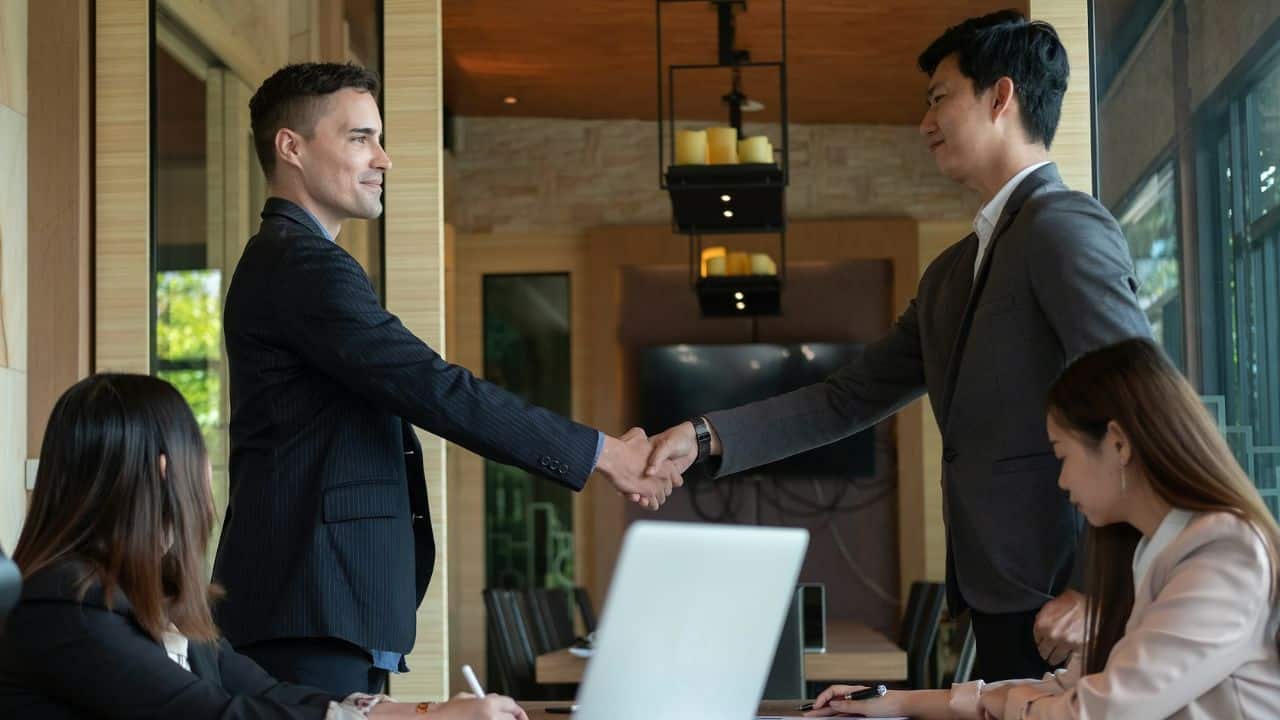 Two diverse businessmen in suits are shaking hands across a conference table.