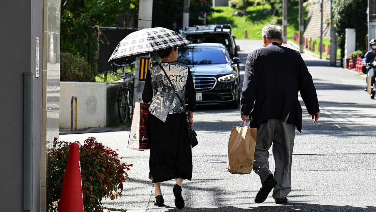A senior couple with shopping bags and an umbrella is walking on a city sidewalk.