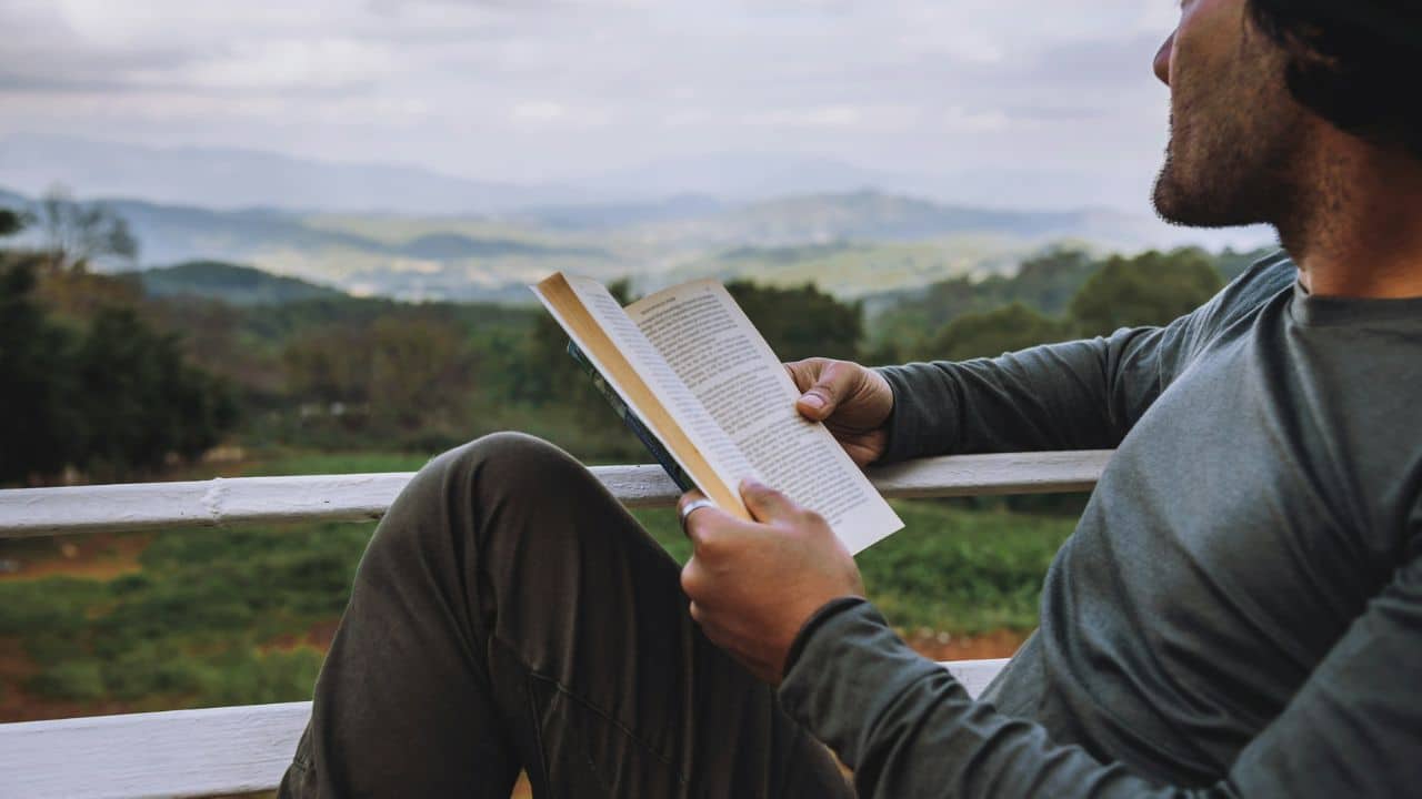 A man is sitting outdoors on a balcony, reading a book.