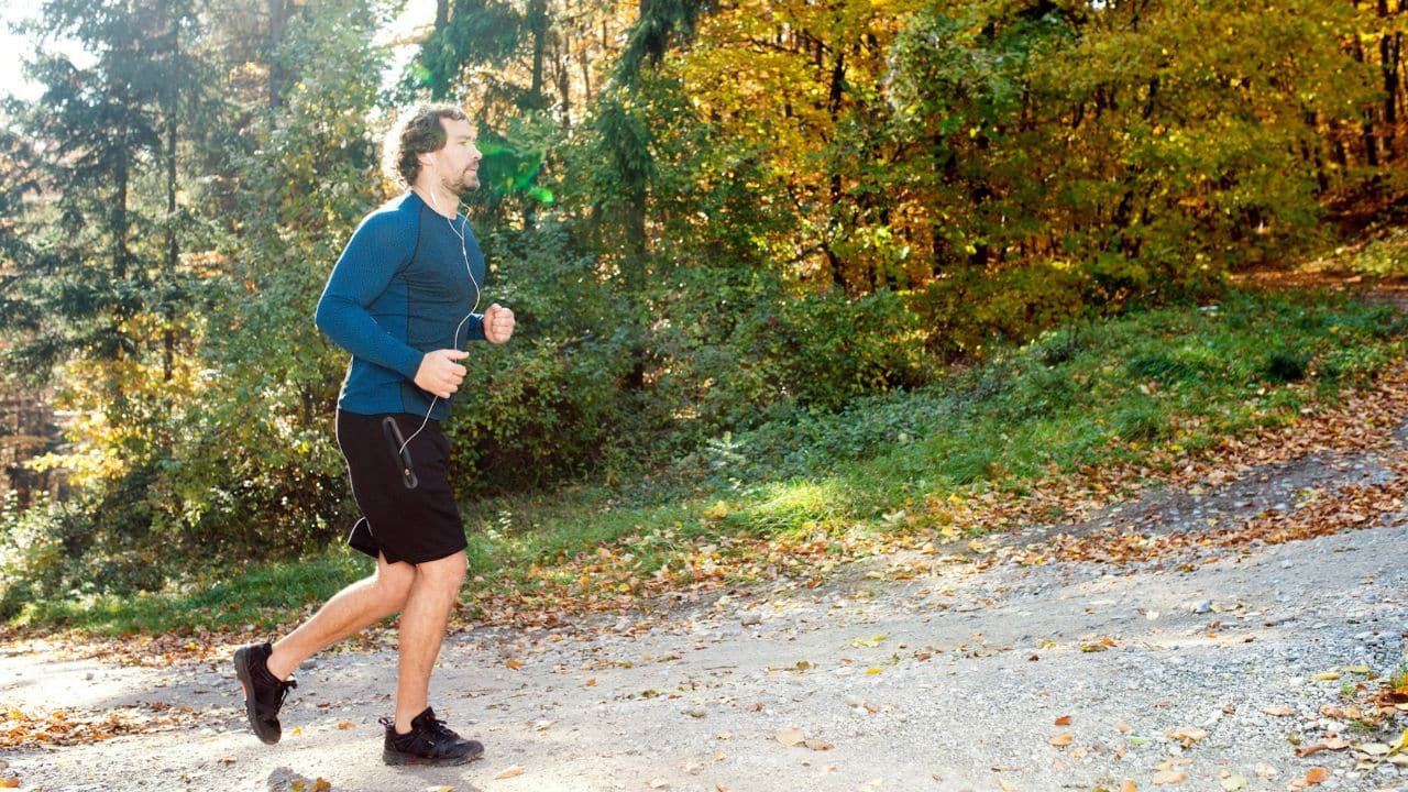 A man with headphones is jogging on a dirt path in a sunlit autumnal forest.