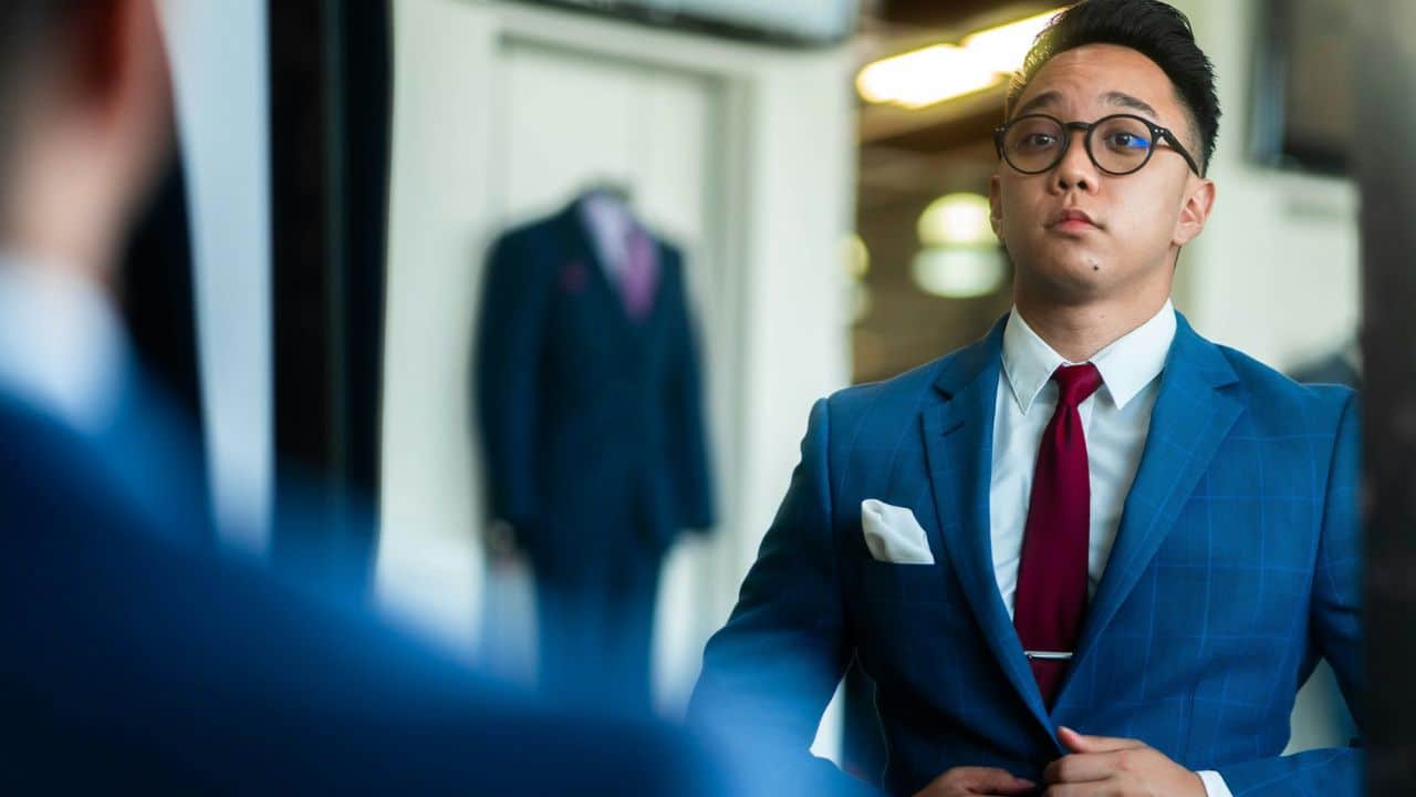 A young man with glasses adjusts his blue suit jacket while looking in a mirror.