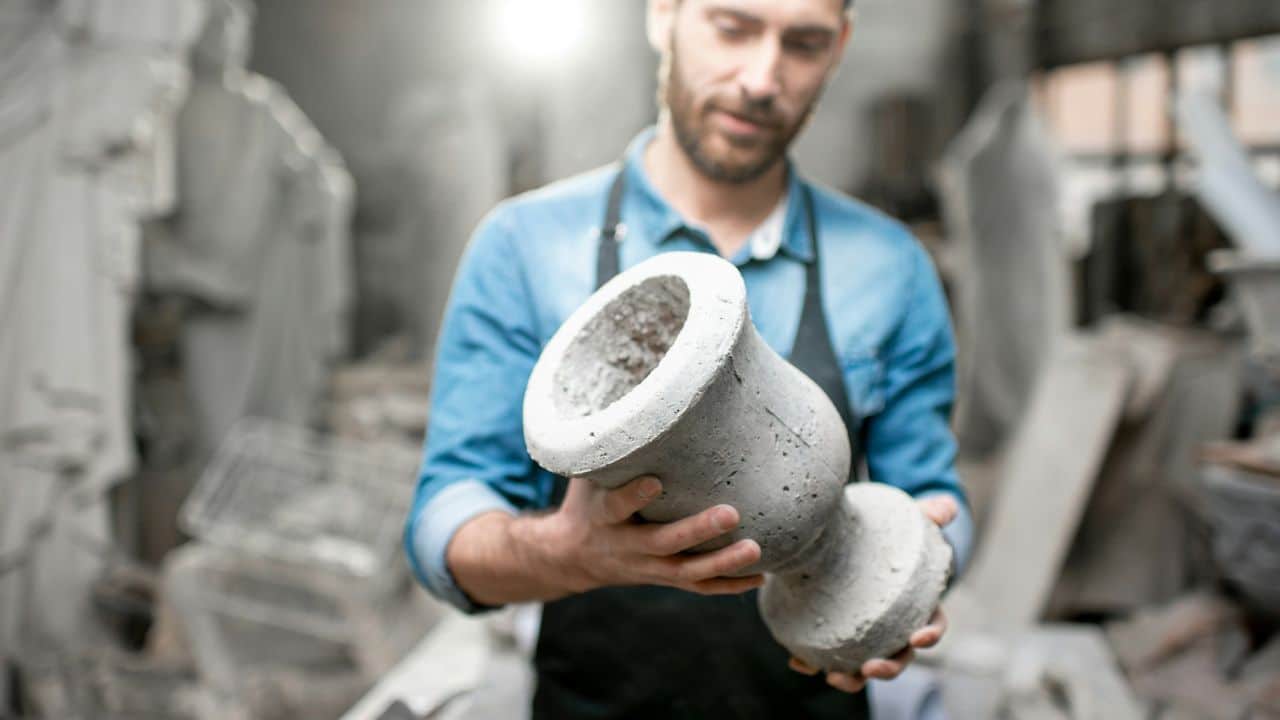 A sculptor in a denim shirt and apron examines a freshly cast concrete vase.