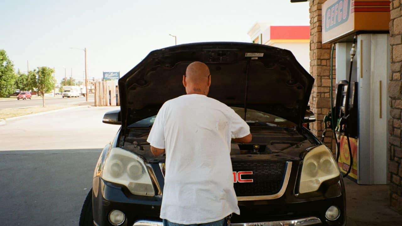A man in a white T-shirt looks under the hood of his black GMC SUV at a gas station.