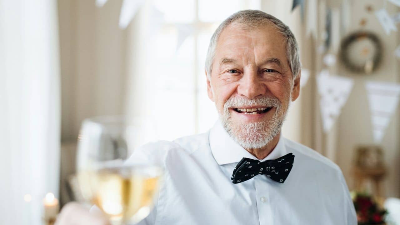 A cheerful, senior man in a white shirt and bow tie holds a glass of wine.